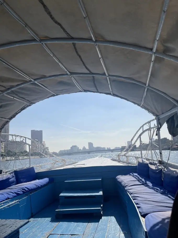 Blick unter dem gewölbten Sonnendach des Boots nach vorn über den Bug, links und rechts blaue Sitzpolster, vorne der Nil, eine Brücke und Hochhäuser