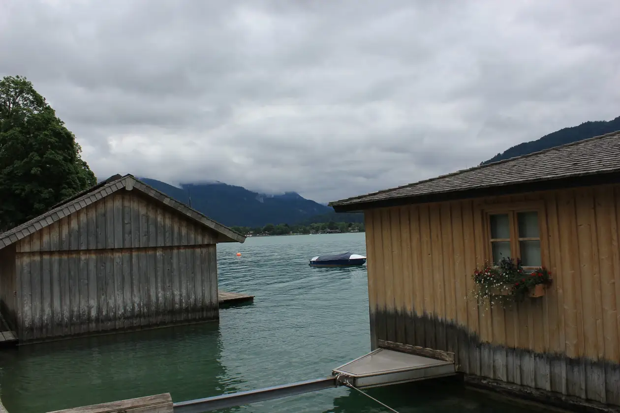 Zwei hölzerne Bootshäuser stehen im Wasser, dazwischen der Blick auf den See mit einem abgedeckten Boot und Bergen im Nebel