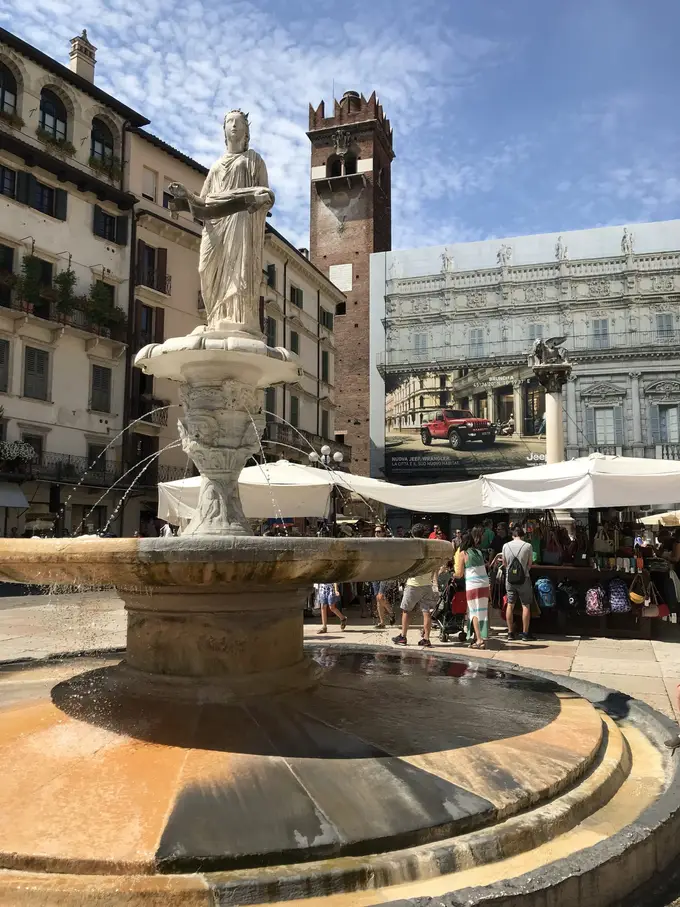 Brunnen mit einer Statue auf der Piazza delle Erbe, dahinter Marktschirme und alte Häuser