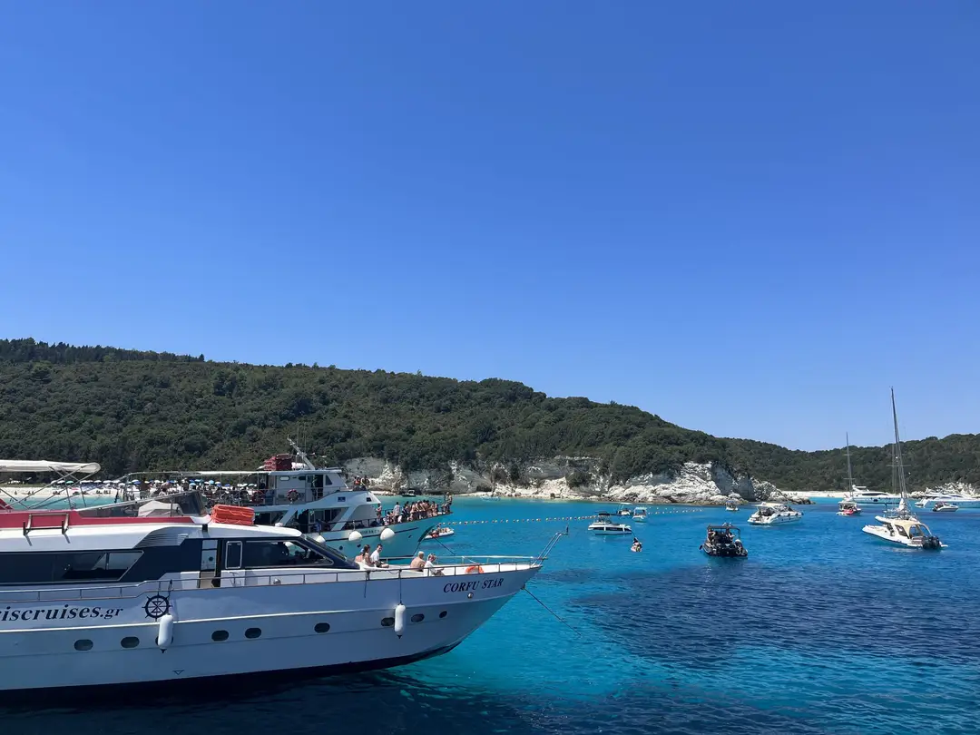 Türkise Bucht vor einem bewaldeten Hügel mit hellem Strand, davor viele Yachten und ein Ausflugsboot