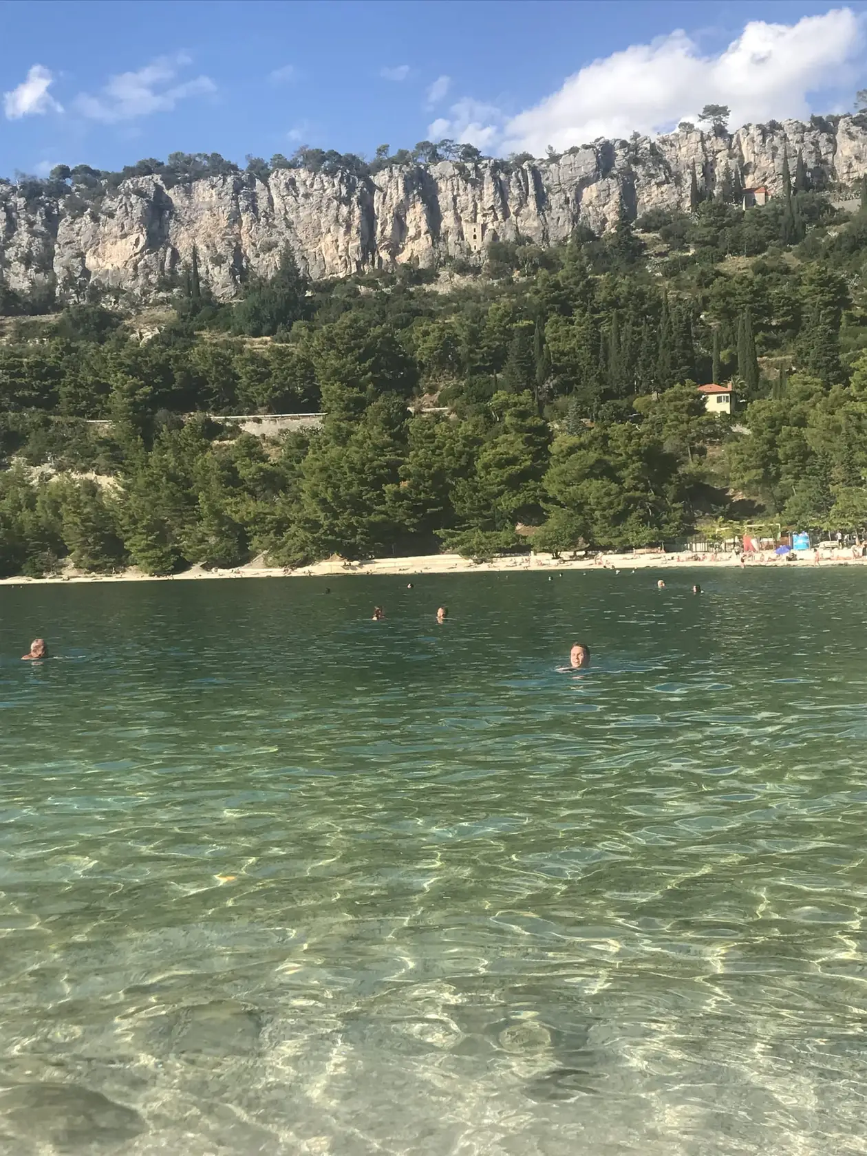 Klares Wasser über hellem Grund, dahinter Schwimmer, ein Kiesstrand, Kiefern und eine Kalkwand
