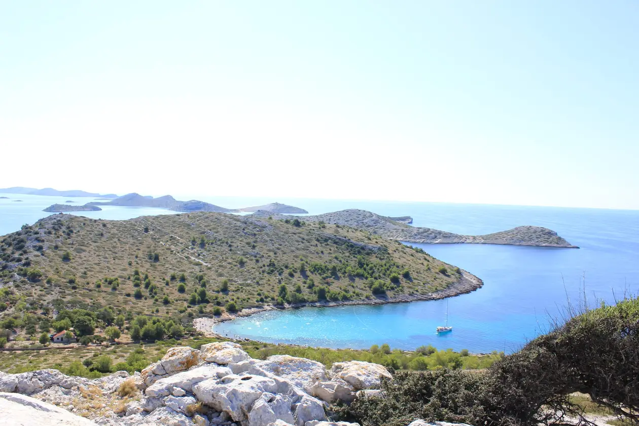 Blick von einer Anhöhe über eine kahle Insel mit einer türkisen Bucht, in der ein Segelboot liegt, dahinter weitere Inseln im Meer