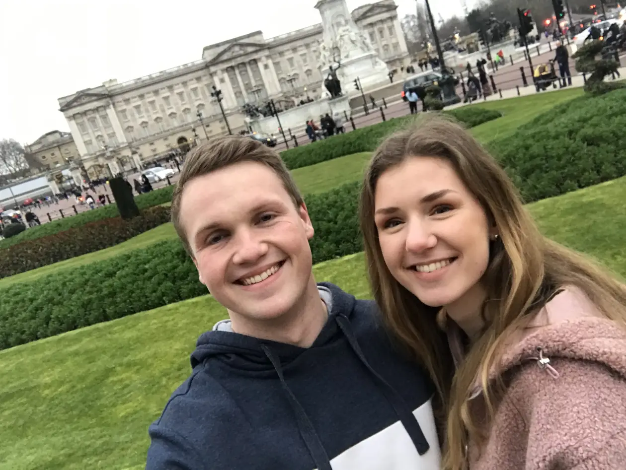 Max und Rebecca lächeln in die Kamera, hinter ihnen Rasen, Hecken und der Buckingham Palace mit dem Victoria Memorial