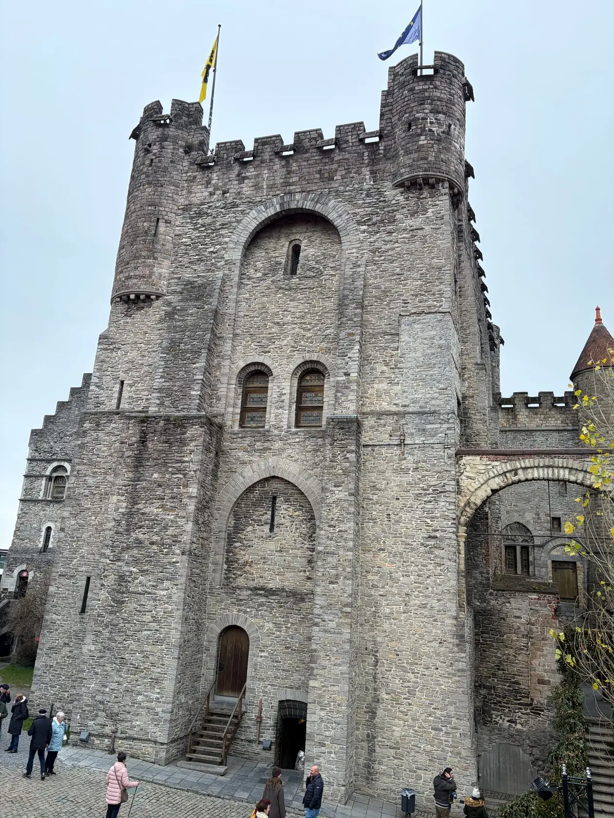 Eine mittelalterliche Burg aus grauem Bruchstein mit Zinnen, zwei runden Ecktürmen und zwei Fahnen, unten auf dem Pflaster einige Besucher