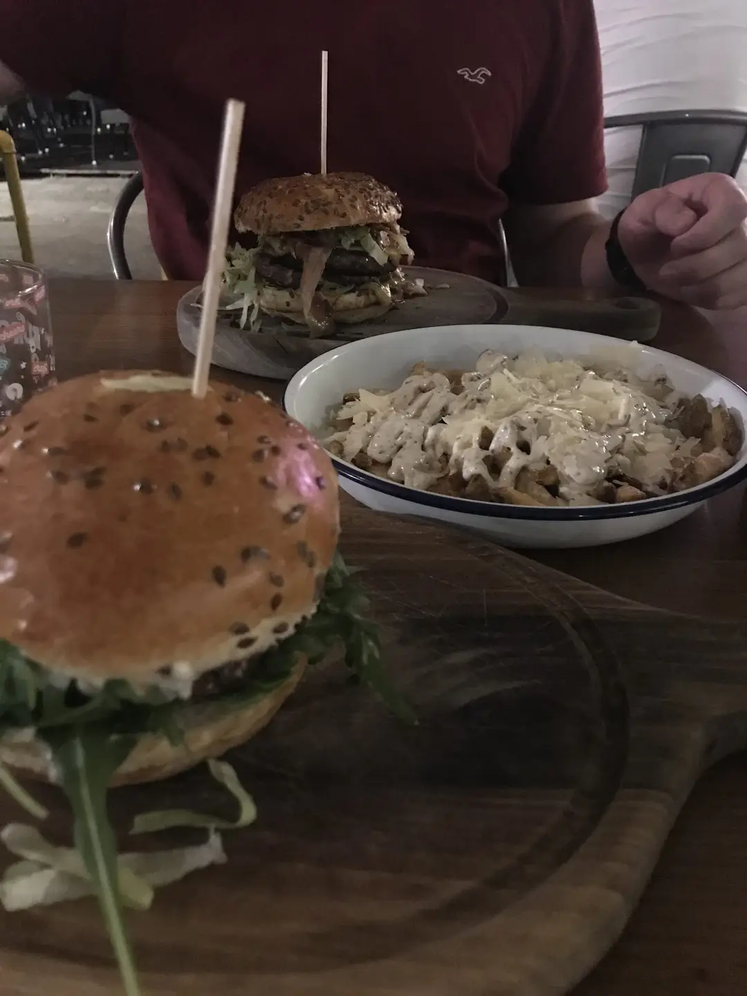 Im dämmrigen Licht zwei Burger auf Holzbrettern und eine Schale Pommes mit heller Soße und gehobeltem Käse, dahinter ein Mann im roten T-Shirt