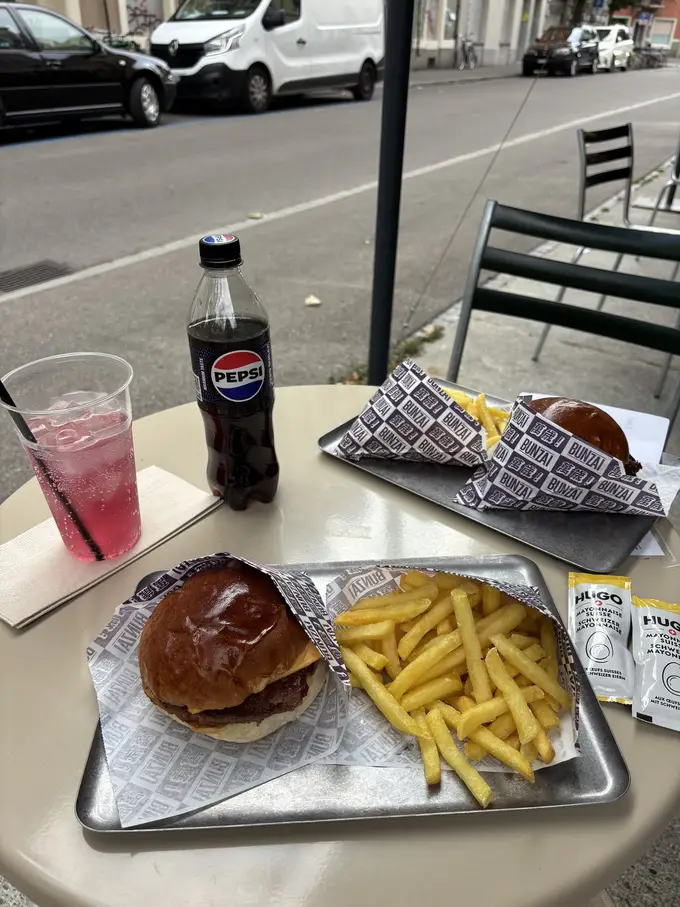 Zwei Burger mit Pommes in bedrucktem Papier auf Metalltabletts, dazu eine Flasche Cola und ein rosa Getränk, auf einem Tisch am Straßenrand