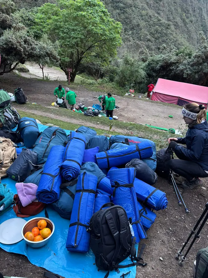 Aufgerollte blaue Schlafsäcke und Rucksäcke auf einer Plane im Camp, dahinter ein rosa Zelt und Männer in grünen Jacken beim Zusammenpacken