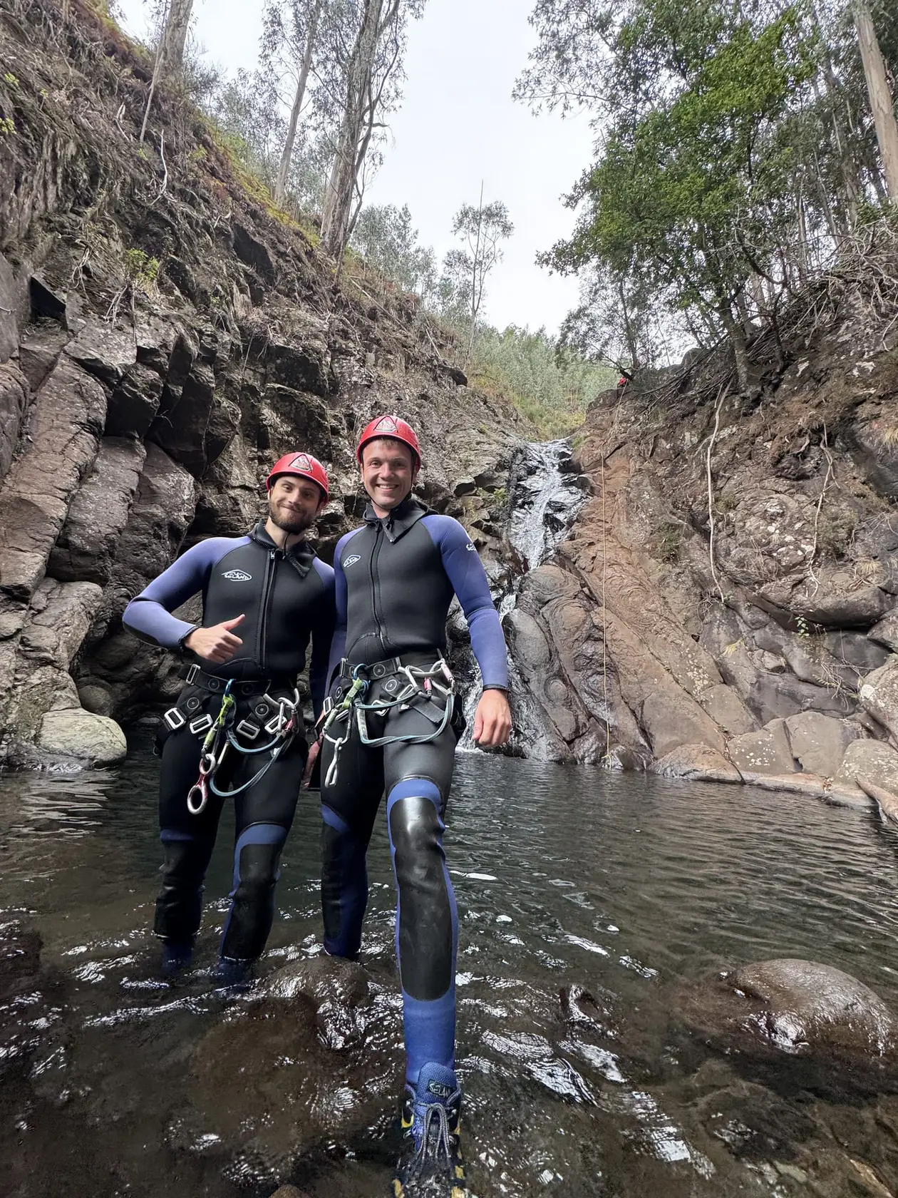 Zwei Männer in Neoprenanzügen und roten Helmen stehen hüfttief im Wasser einer bewaldeten Schlucht und lächeln