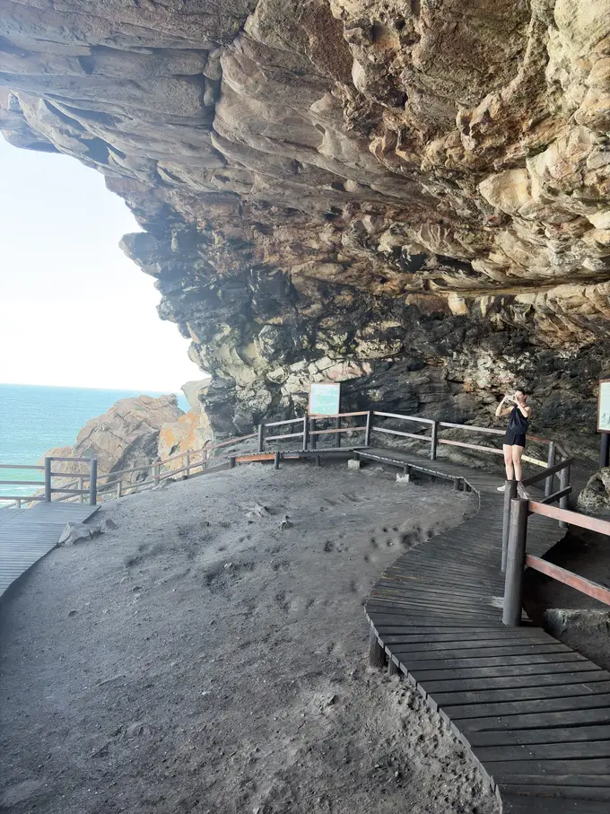 Holzsteg führt zu einer Aussichtsplattform vor einer Felshöhle über dem Meer, eine Person steht am Geländer