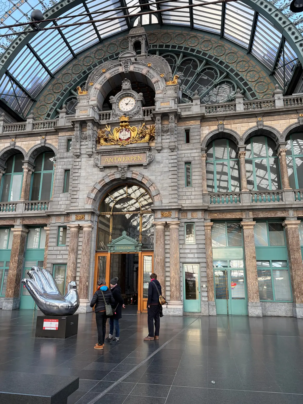Innenwand der Bahnhofshalle von Antwerpen-Centraal mit Uhr, vergoldetem Wappen und dem Schriftzug Antwerpen unter einem Glasdach, links eine silberne Skulptur einer Hand