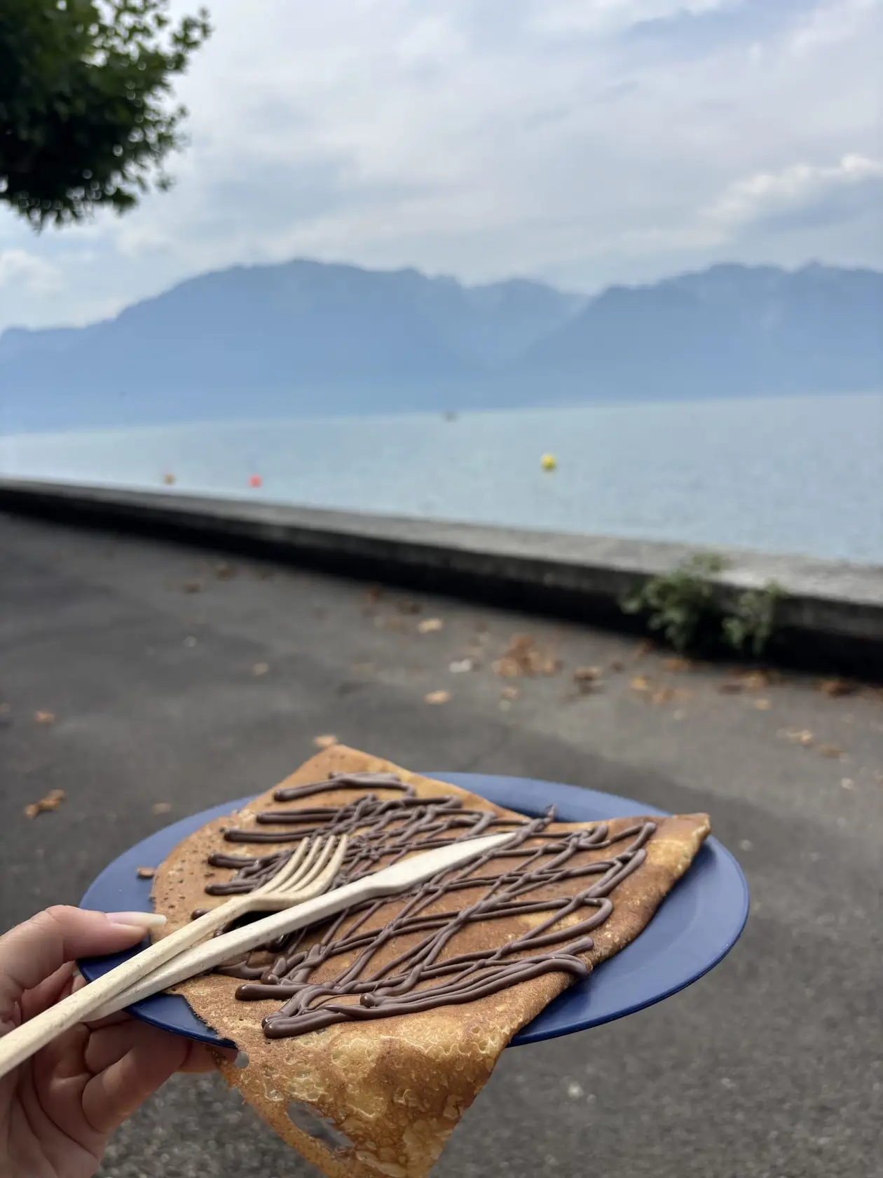 Eine Hand hält einen Teller mit Crêpe und Schokoladensauce, dahinter eine Ufermauer, der Genfersee und Berge im Dunst