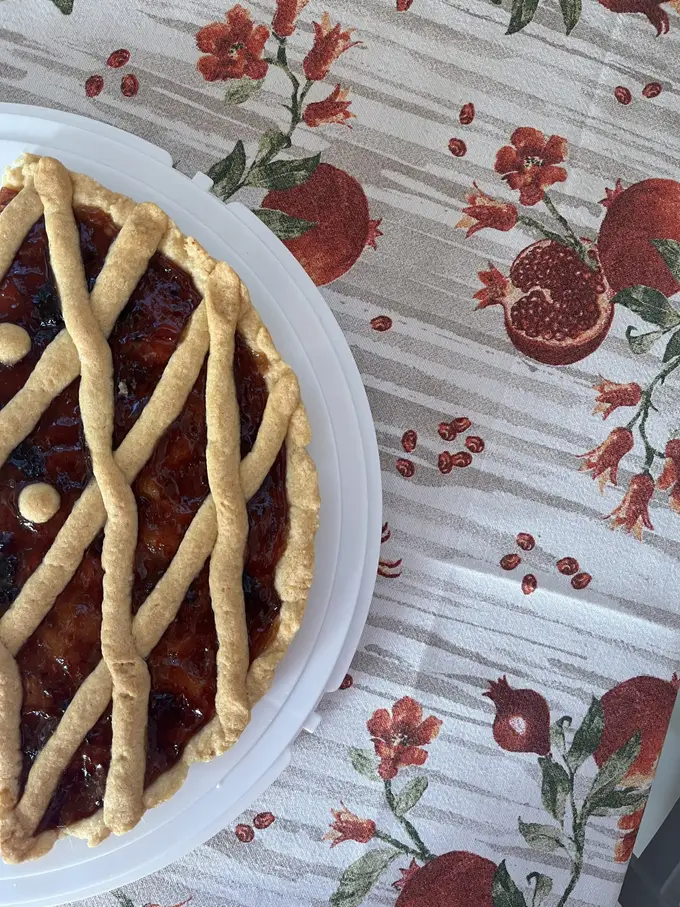 Ein Gitterkuchen mit dunkler Marmeladenfüllung auf einem weißen Teller, darunter eine Tischdecke mit Granatäpfeln und Blüten