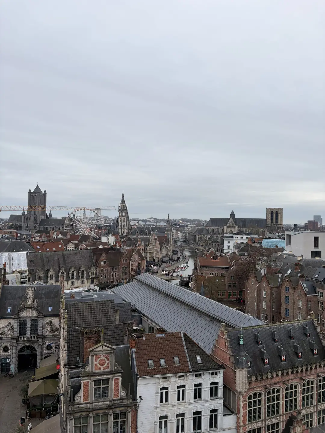 Blick von oben über die Dächer der Genter Altstadt auf die Leie, an deren Ufer sich Giebelhäuser reihen, dahinter Kirchtürme und ein Riesenrad unter grauem Himmel
