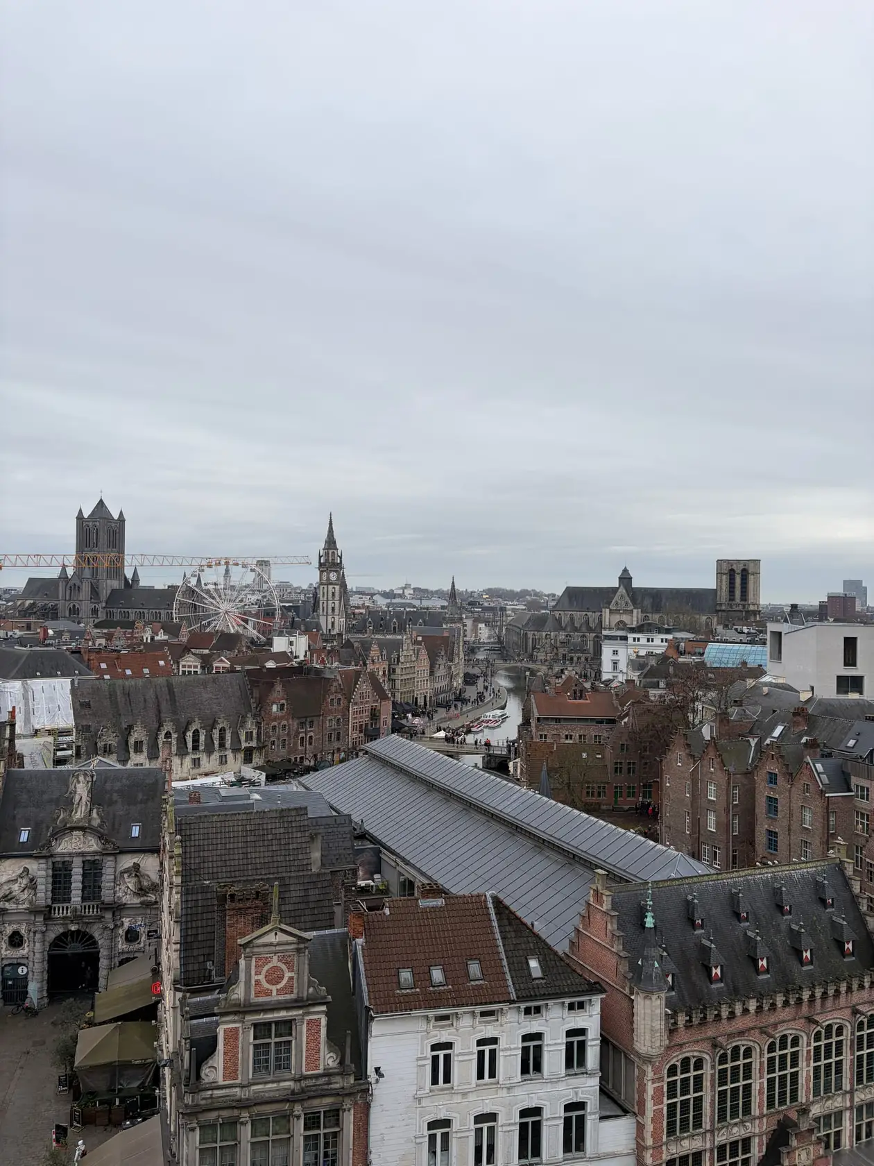 Blick von oben über die Dächer der Genter Altstadt auf die Leie, an deren Ufer sich Giebelhäuser reihen, dahinter Kirchtürme und ein Riesenrad unter grauem Himmel