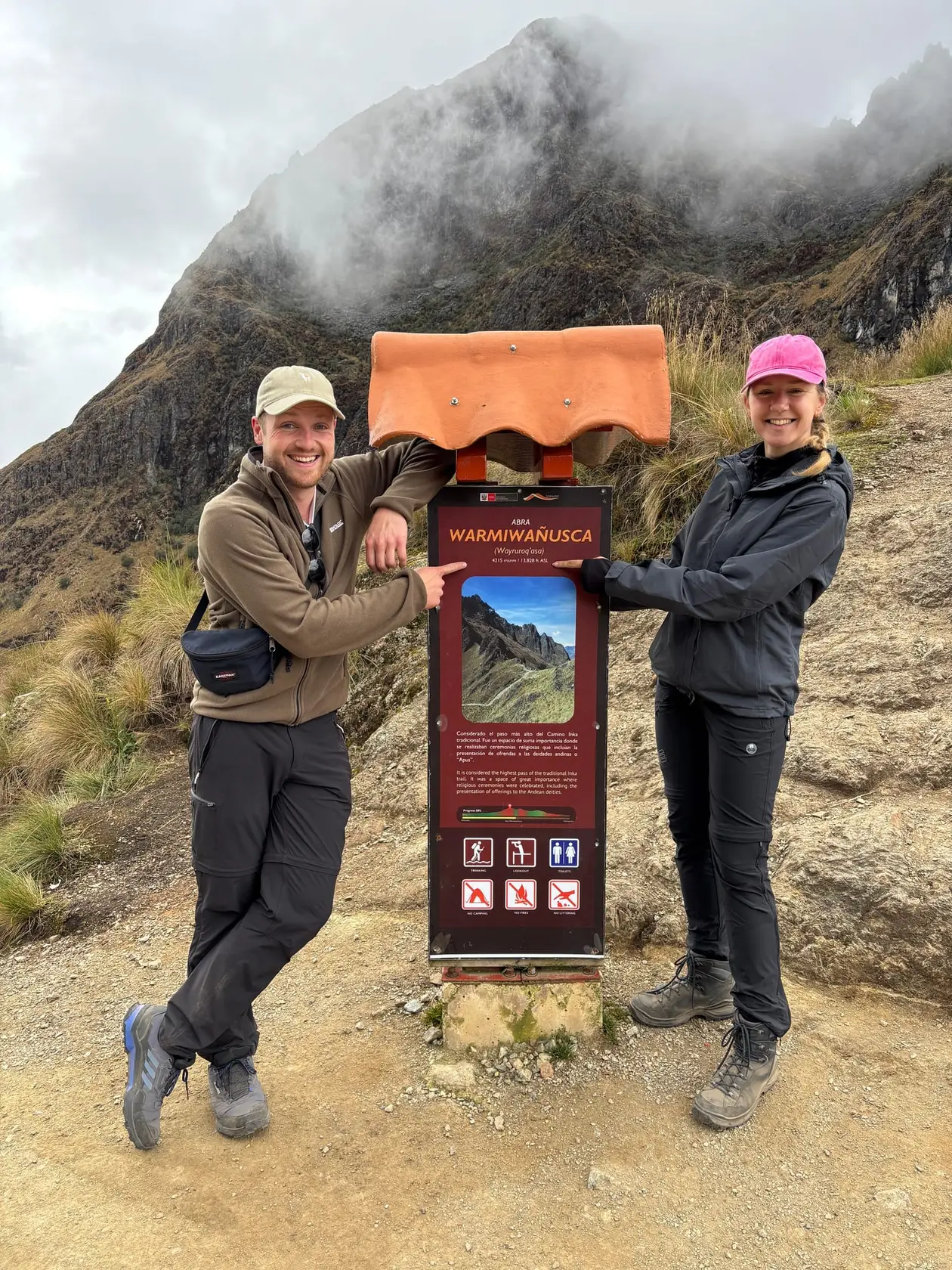 Max und Rebecca stehen links und rechts vom Schild „Abra Warmiwañusca“ auf dem Pass, dahinter ein wolkenverhangener Felsgipfel