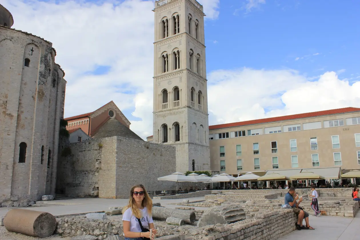 Rebecca mit Sonnenbrille vor den Mauerresten des Forums, links die runde Donatuskirche, dahinter der Glockenturm