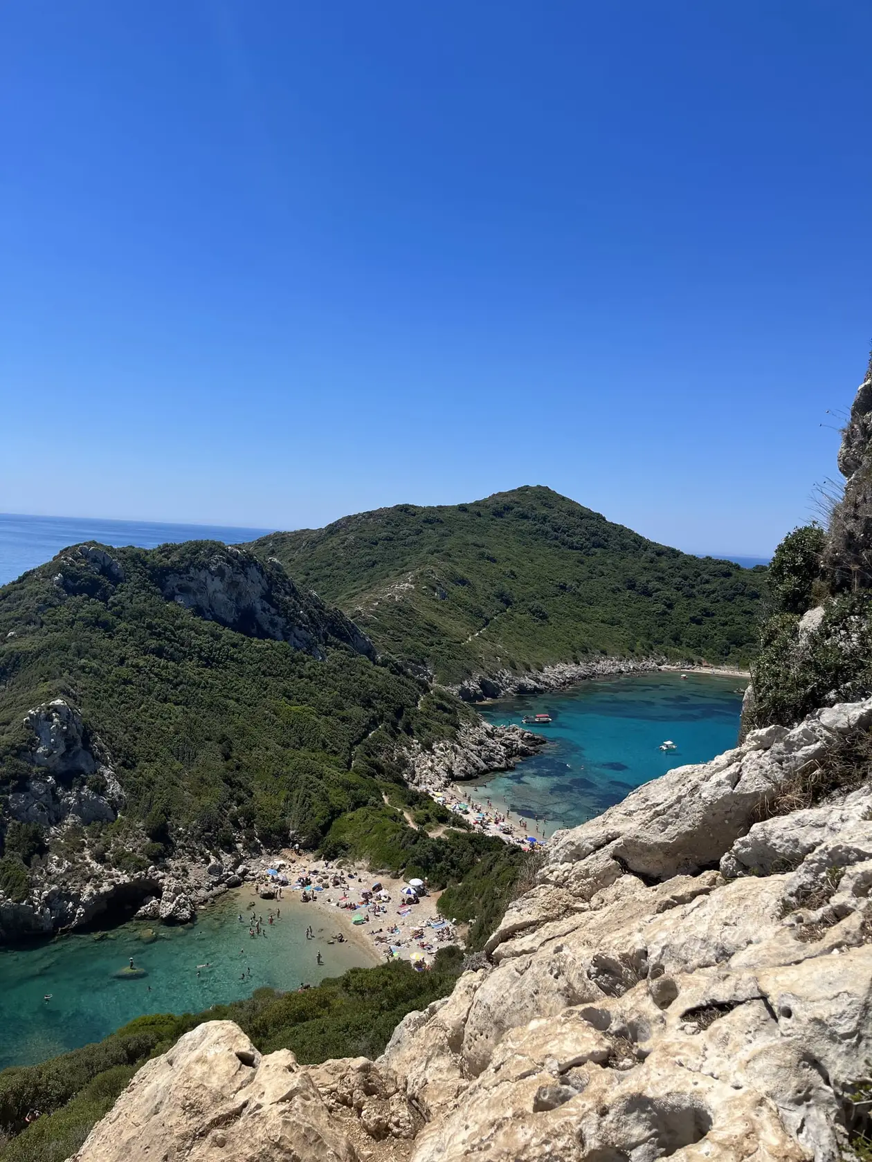 Blick von einem Felsen hinunter auf zwei Buchten mit türkisem Wasser, getrennt von einer schmalen bewachsenen Landbrücke