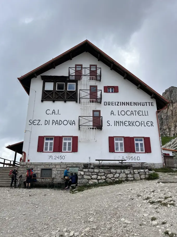 Die weiße Dreizinnenhütte mit roten Fensterläden und der Aufschrift „Dreizinnenhütte“ und „M. 2450“, davor Wanderer an Holztischen