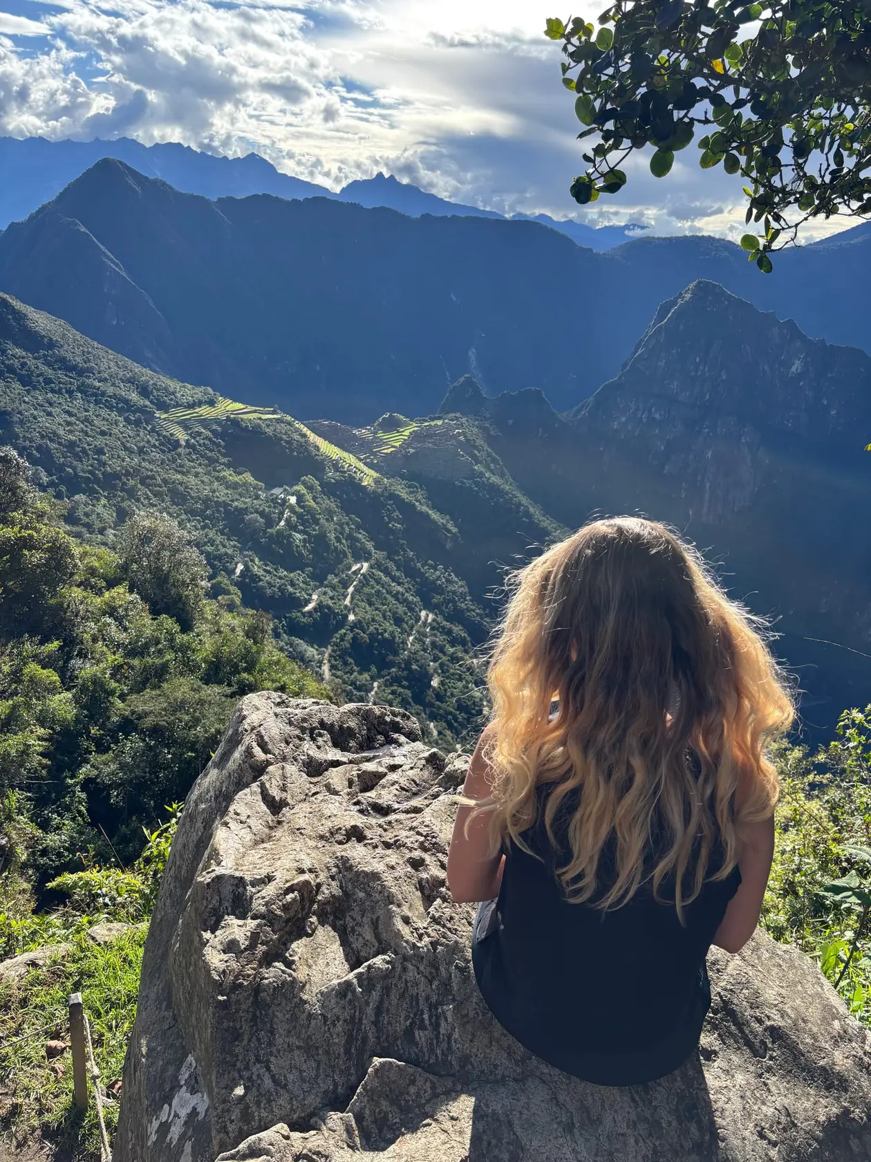 Rebecca sitzt von hinten gesehen auf einem Felsen und schaut über das Tal auf die ferne Ruinenstadt Machu Picchu im Nachmittagslicht