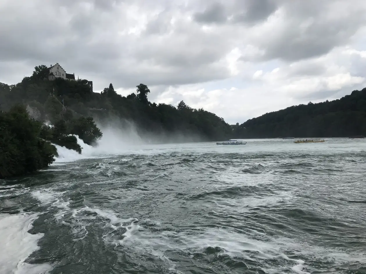 Aufgewühltes Wasser unter dem Rheinfall, links stürzt das Wasser mit viel Gischt zwischen Bäumen herab, darüber auf dem Hang ein Schloss, auf dem Fluss zwei Ausflugsboote