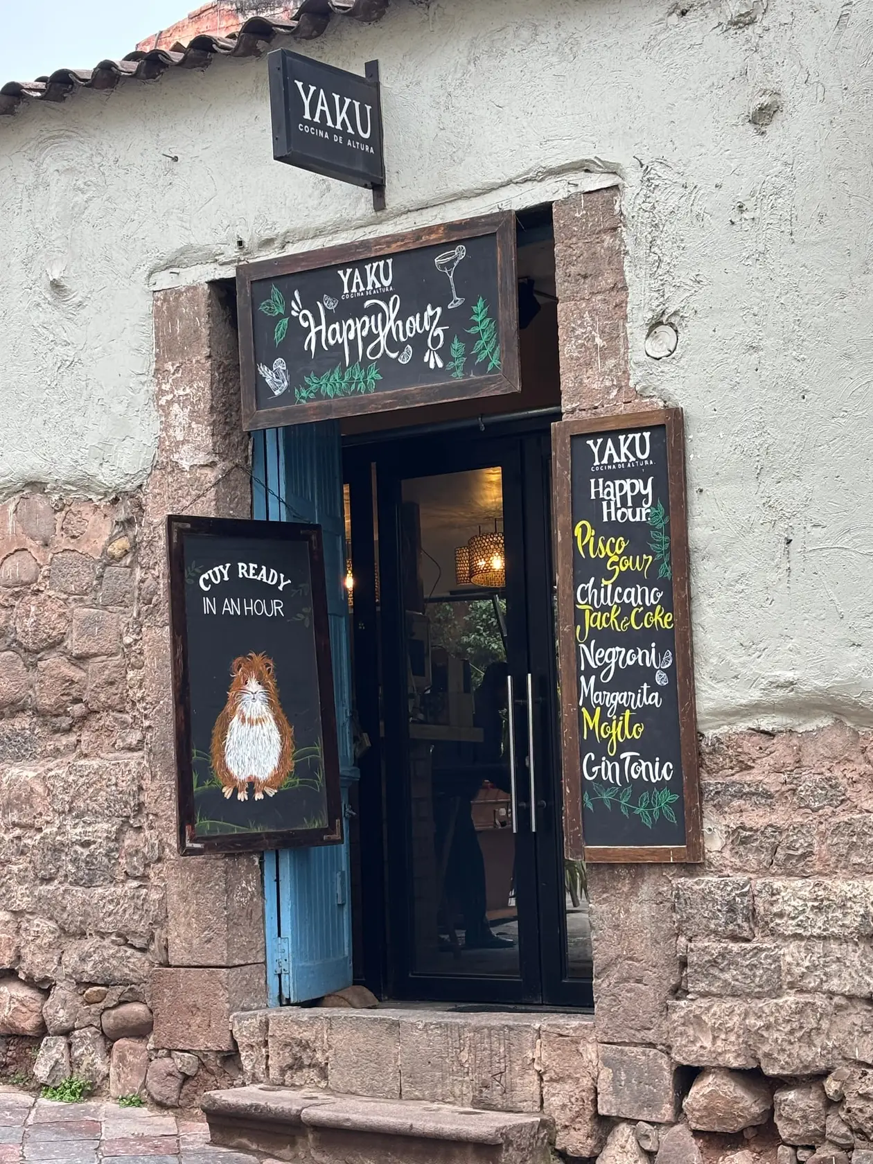 Eingang einer Bar in einer Steinmauer, daneben Kreidetafeln mit Happy-Hour-Cocktails und dem Hinweis „Cuy ready in an hour“ mit gezeichnetem Meerschweinchen