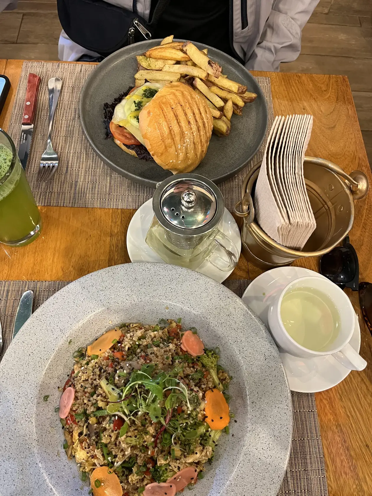 Tisch mit einem bunten Salat mit Getreide und Karotten und einem Burger mit Pommes, dazu Tee und ein grüner Saft