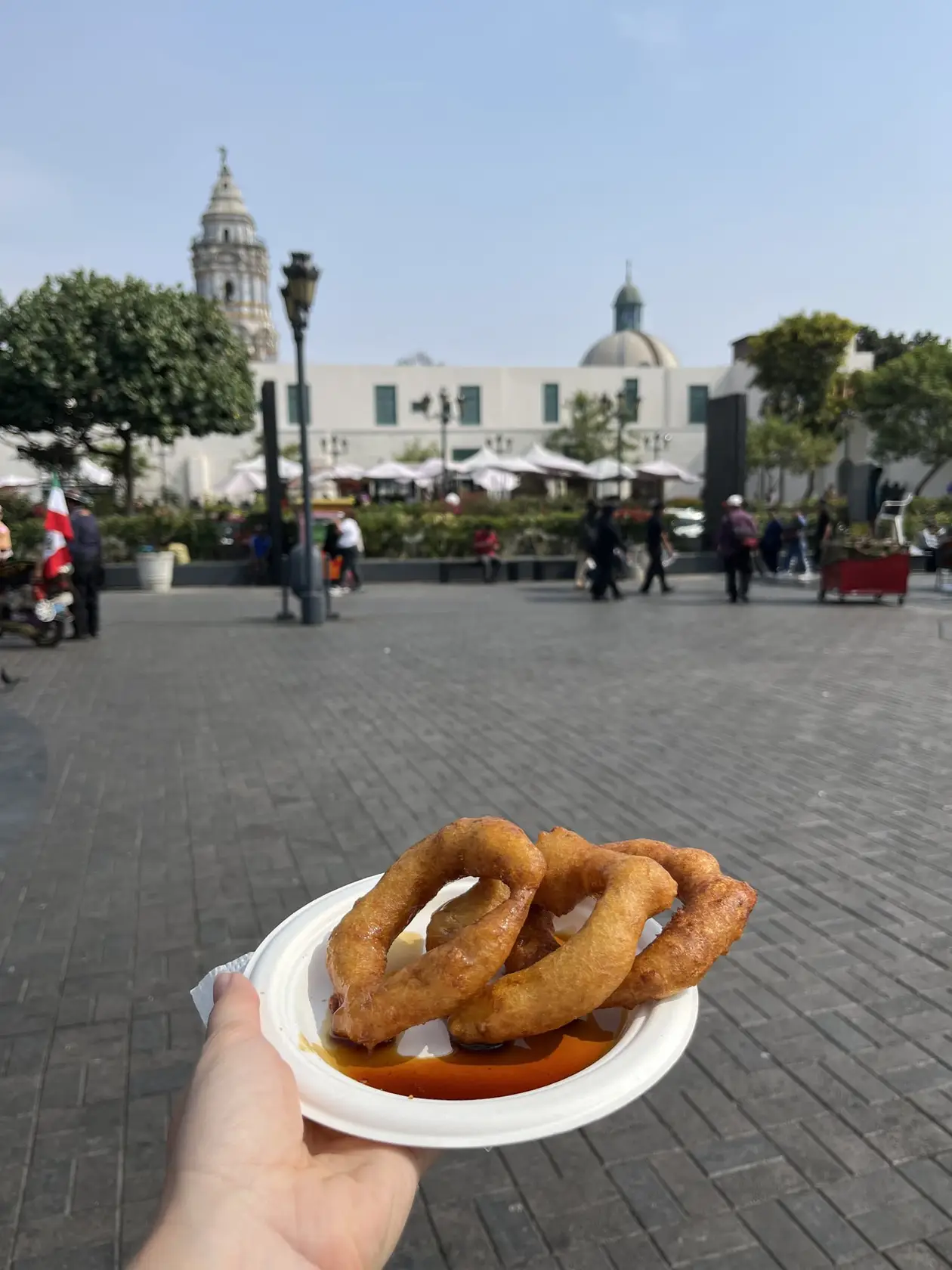 Eine Hand hält einen Pappteller mit ringförmigen frittierten Picarones in Sirup, dahinter die Plaza de Armas