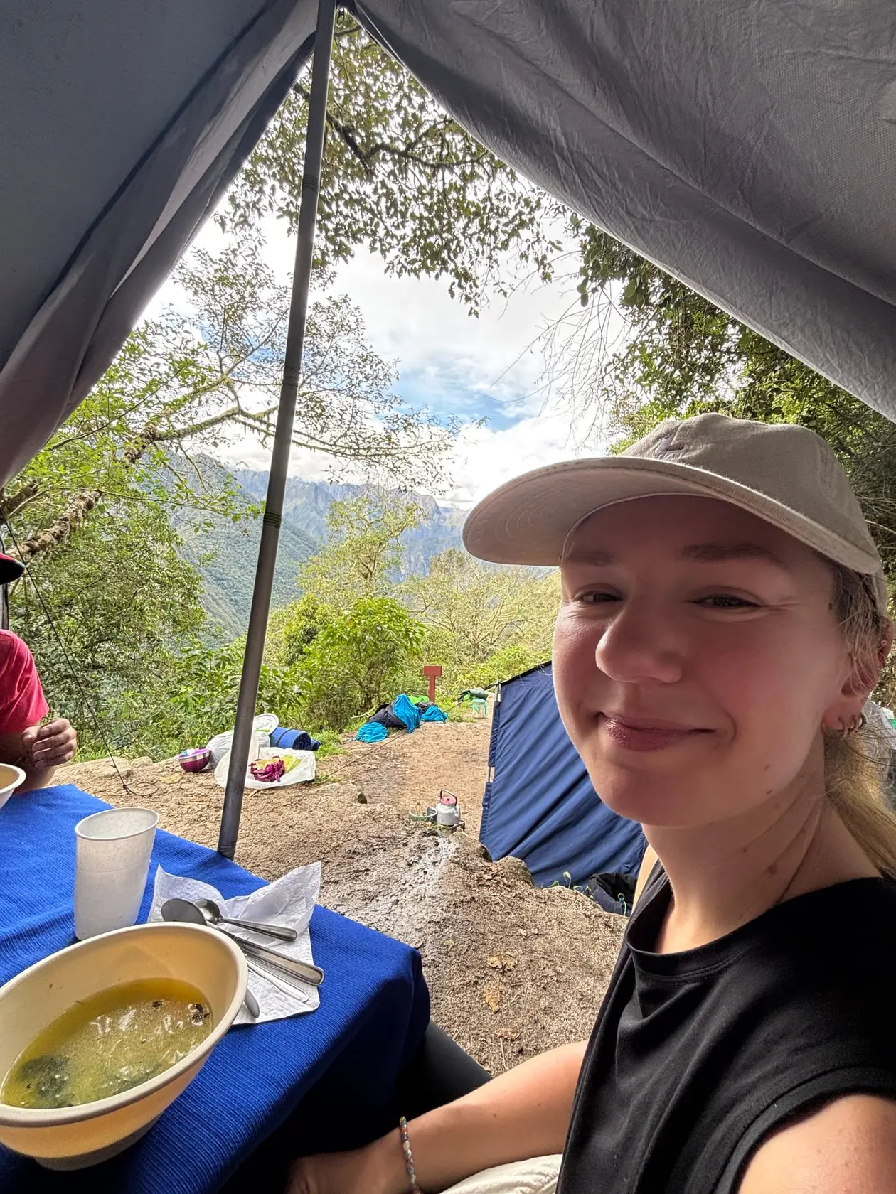 Rebecca lächelt im Essenszelt, vor ihr ein Teller Suppe auf blauer Tischdecke, draußen Bäume und Berge