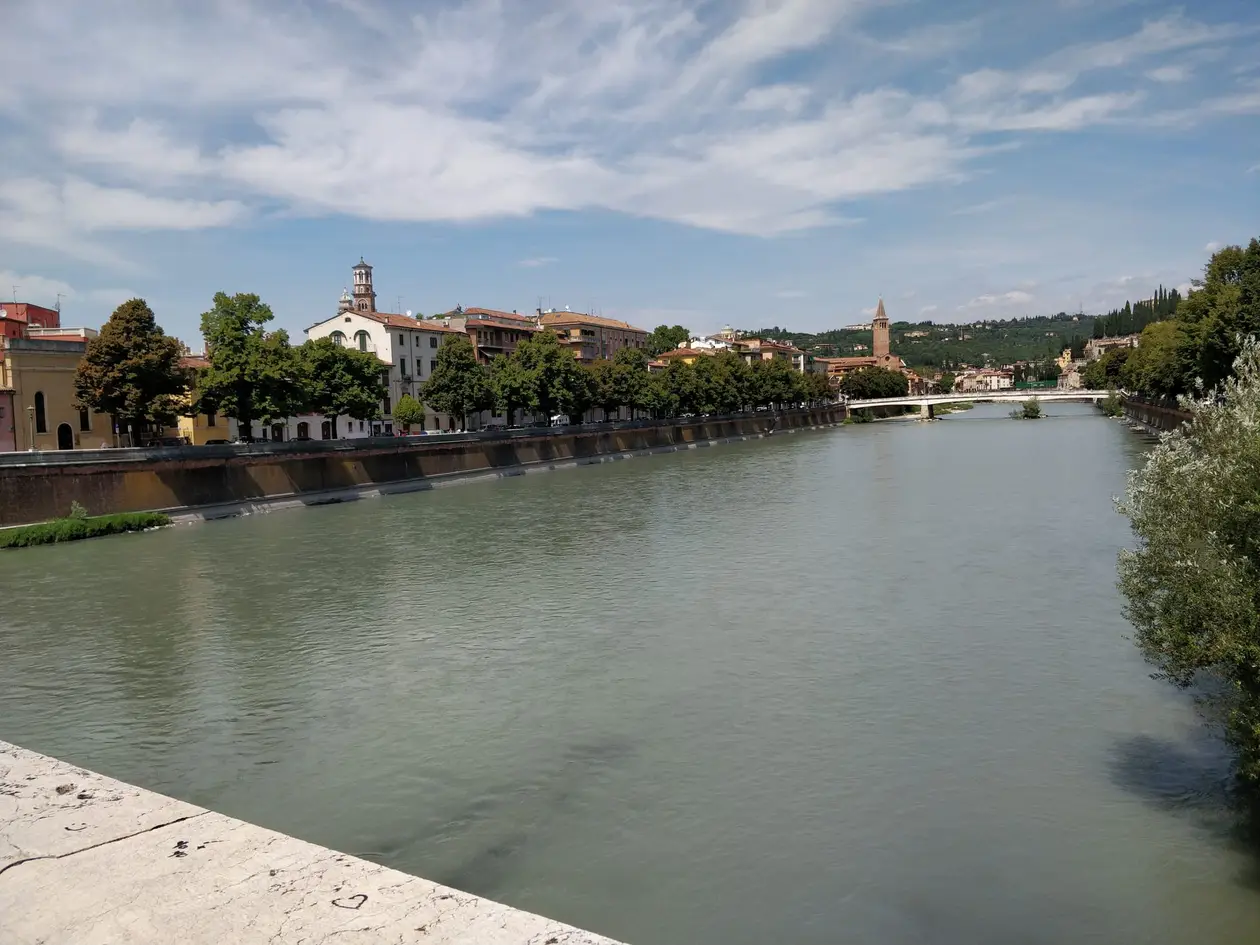 Die breite Etsch zwischen Uferbäumen, am Horizont Häuser, ein Kirchturm und eine Brücke