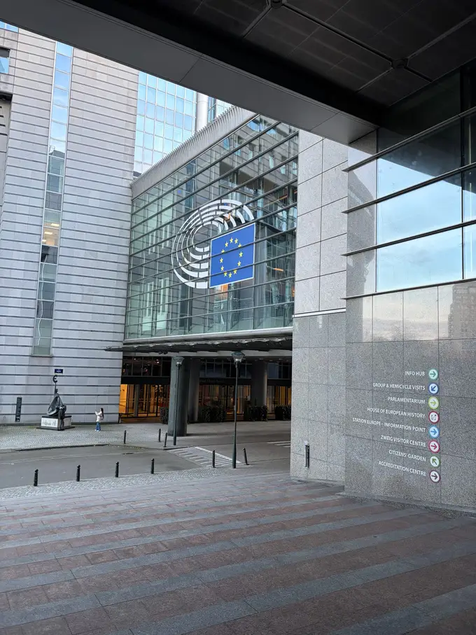 Eingang des Europäischen Parlaments mit dem Parlamentslogo und der Europaflagge an einer Glasfassade, rechts Wegweiser zu den Besucherbereichen