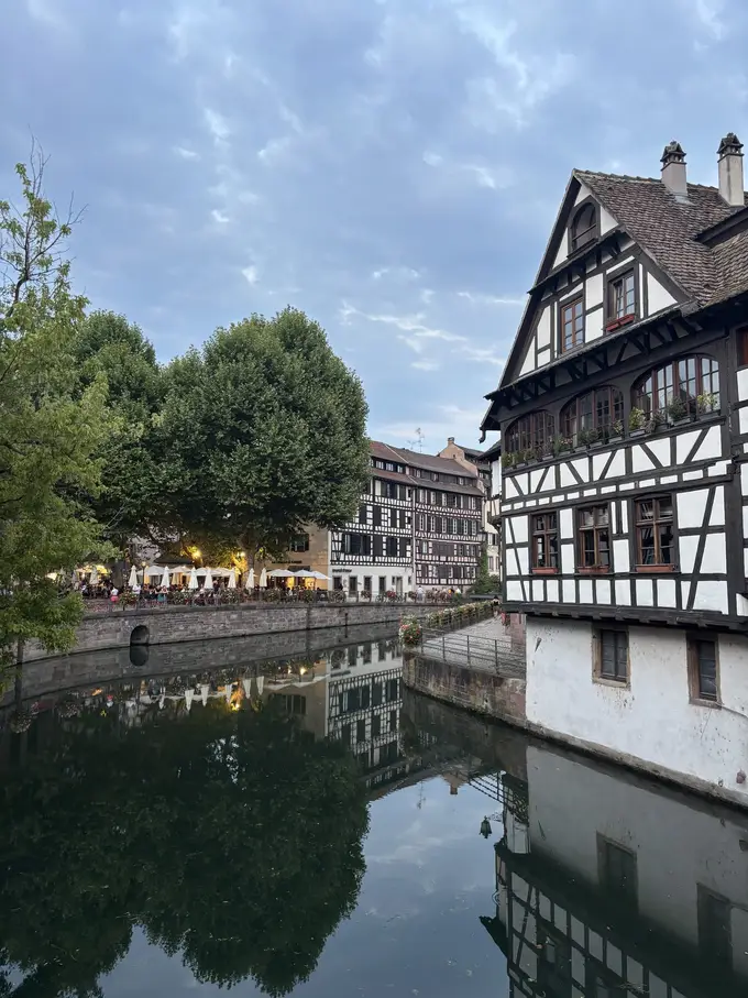 Ein großes schwarz-weißes Fachwerkhaus direkt am Kanal, das sich im Wasser spiegelt, links Platanen und eine beleuchtete Terrasse