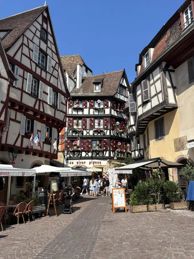 Eine gepflasterte Gasse zwischen Fachwerkhäusern, am Ende ein Haus mit bunten Herzen an den Fensterläden, davor Restauranttische und Passanten