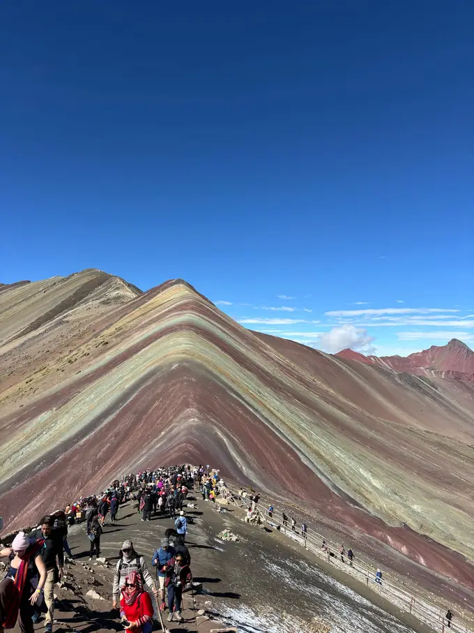 Die farbigen Gesteinsschichten des Rainbow Mountain in Rot, Grün, Ocker und Violett, unten eine Menschenmenge auf dem Aussichtsgrat
