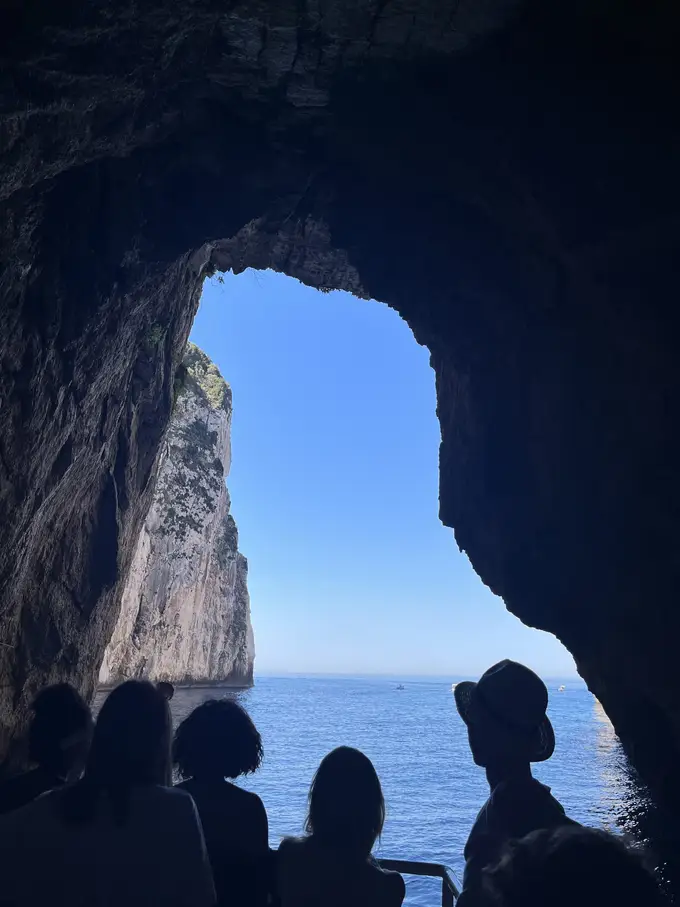 Blick aus einer dunklen Höhle durch ein Felsentor aufs Meer, davor die Silhouetten von Passagieren