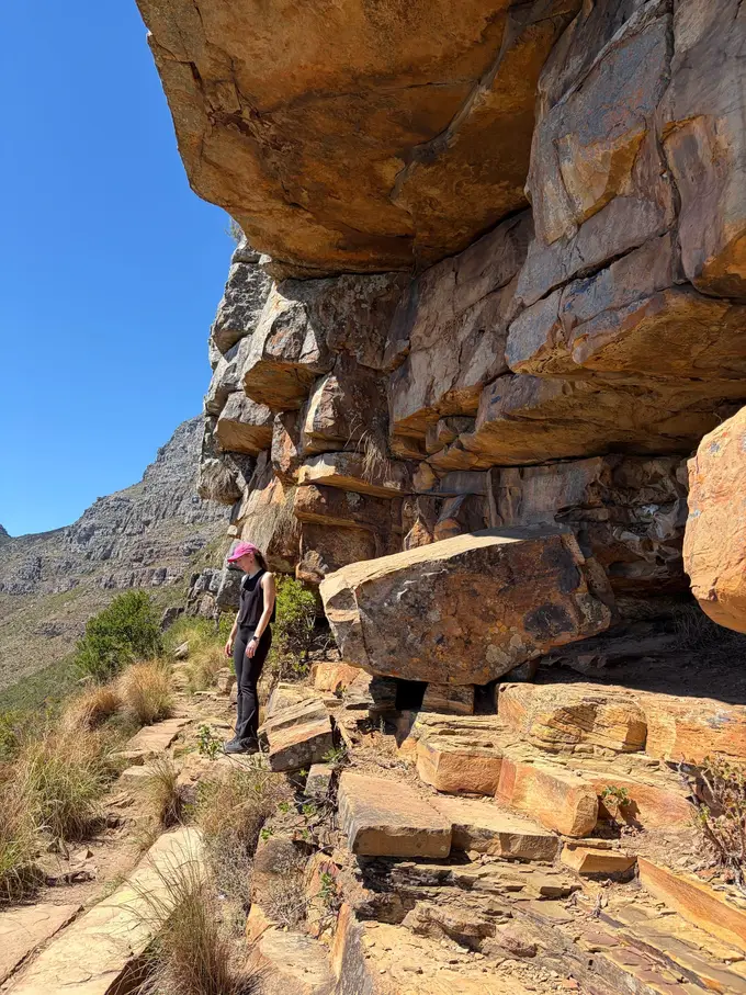 Rebecca steht zwischen mächtigen geschichteten Sandsteinfelsen am Wegrand auf dem Tafelberg