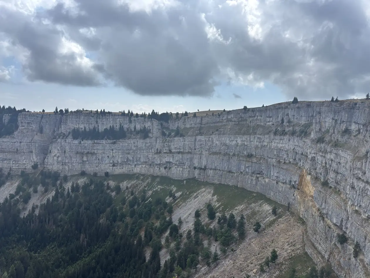 Die halbrunde, fast senkrechte Felswand des Creux du Van, am Fuß bewaldete Geröllhänge, darüber Wolken
