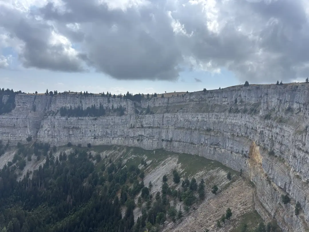 Die halbrunde, fast senkrechte Felswand des Creux du Van, am Fuß bewaldete Geröllhänge, darüber Wolken