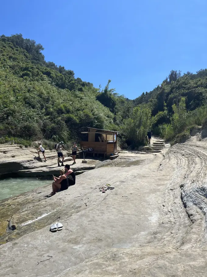 Flache Felsplatten am Wasser unter einem bewaldeten Hang, hinten eine kleine Holzhütte und ein paar Menschen