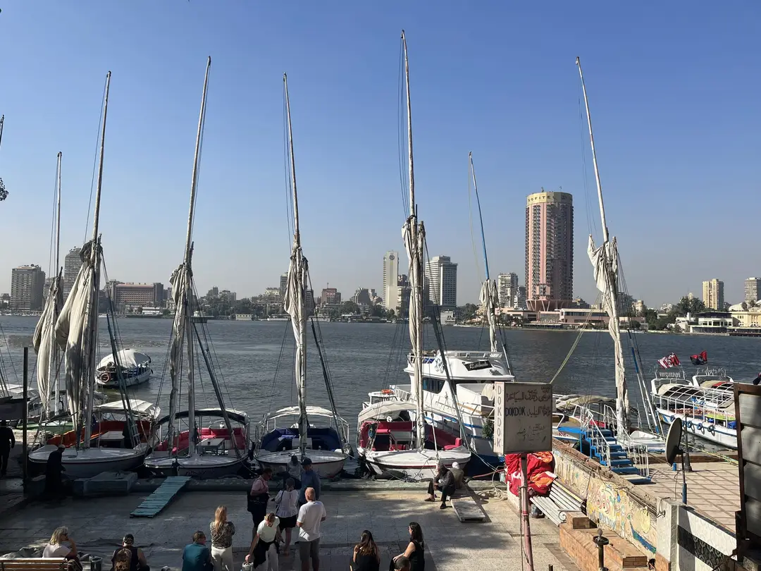 Mehrere Segelboote mit eingerollten Segeln liegen am Nilufer, davor stehen Menschen an der Anlegestelle, am anderen Ufer Hochhäuser
