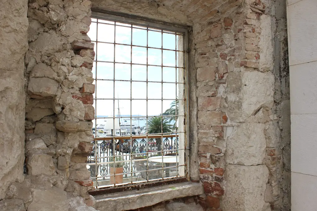 Ein vergittertes Fenster in einer bröckelnden Mauer aus Stein und Ziegeln, draußen Palmen, Boote und der Hafen