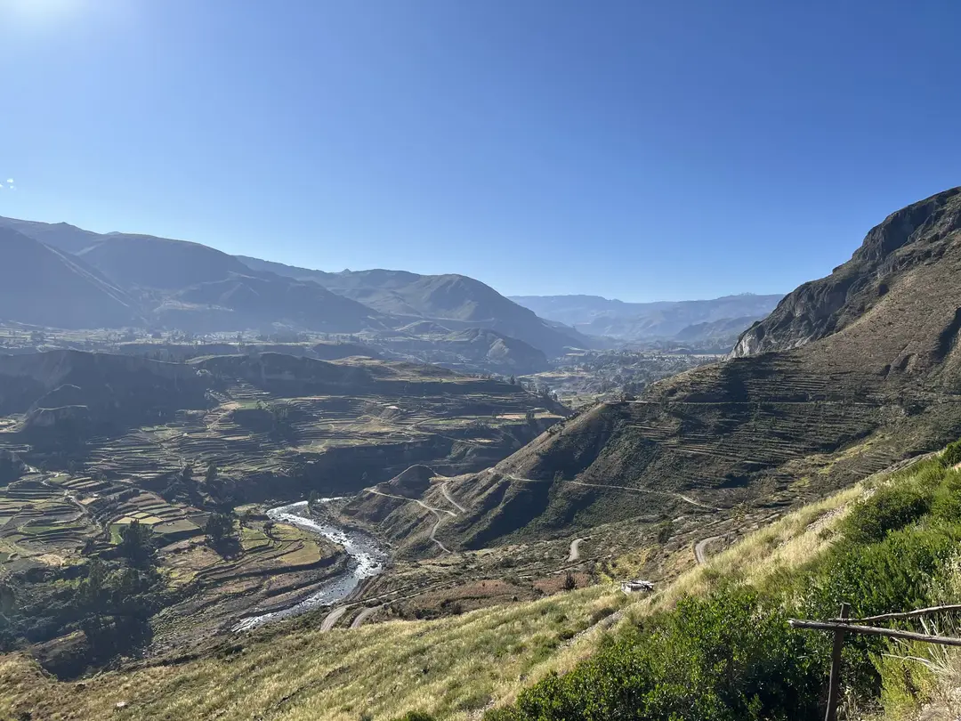 Der Colca-Fluss windet sich durch ein Tal mit Ackerterrassen an den Hängen, dahinter Bergketten unter blauem Himmel