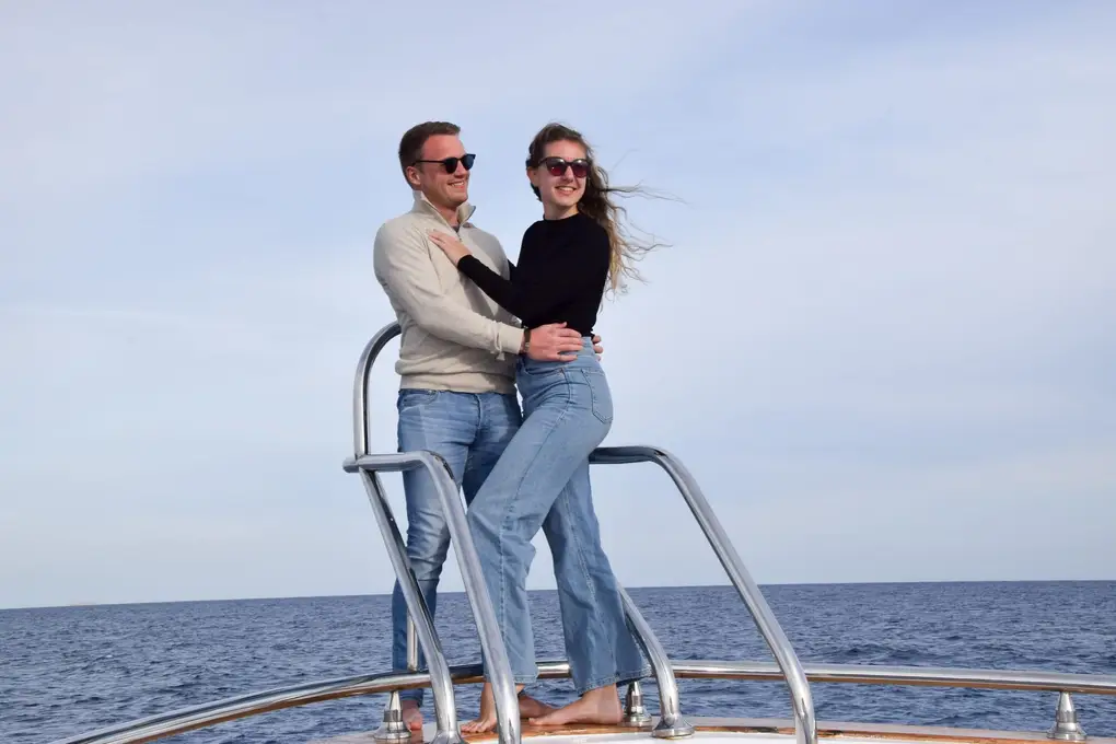 Max und Rebecca stehen umarmt am Bug des Boots zwischen den Relingstangen, hinter ihnen offenes Meer
