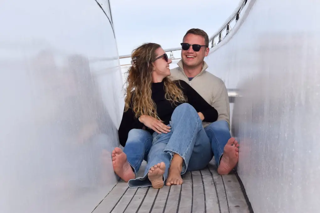 Max und Rebecca sitzen barfuß und lachend in einem schmalen Durchgang an Deck, eng aneinander