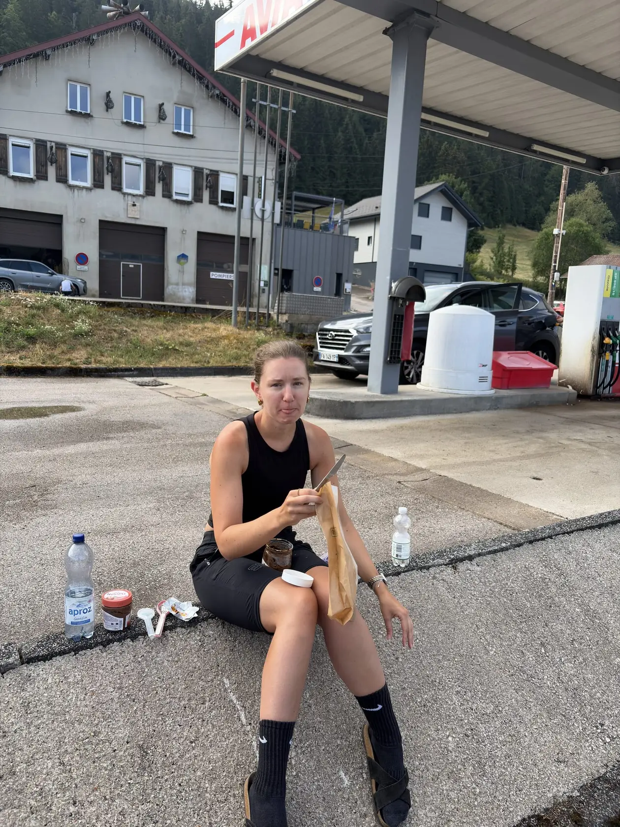 Rebecca sitzt auf einem Bordstein und isst ein belegtes Brötchen, neben ihr Wasserflaschen und ein Glas Aufstrich, dahinter Zapfsäulen und Häuser