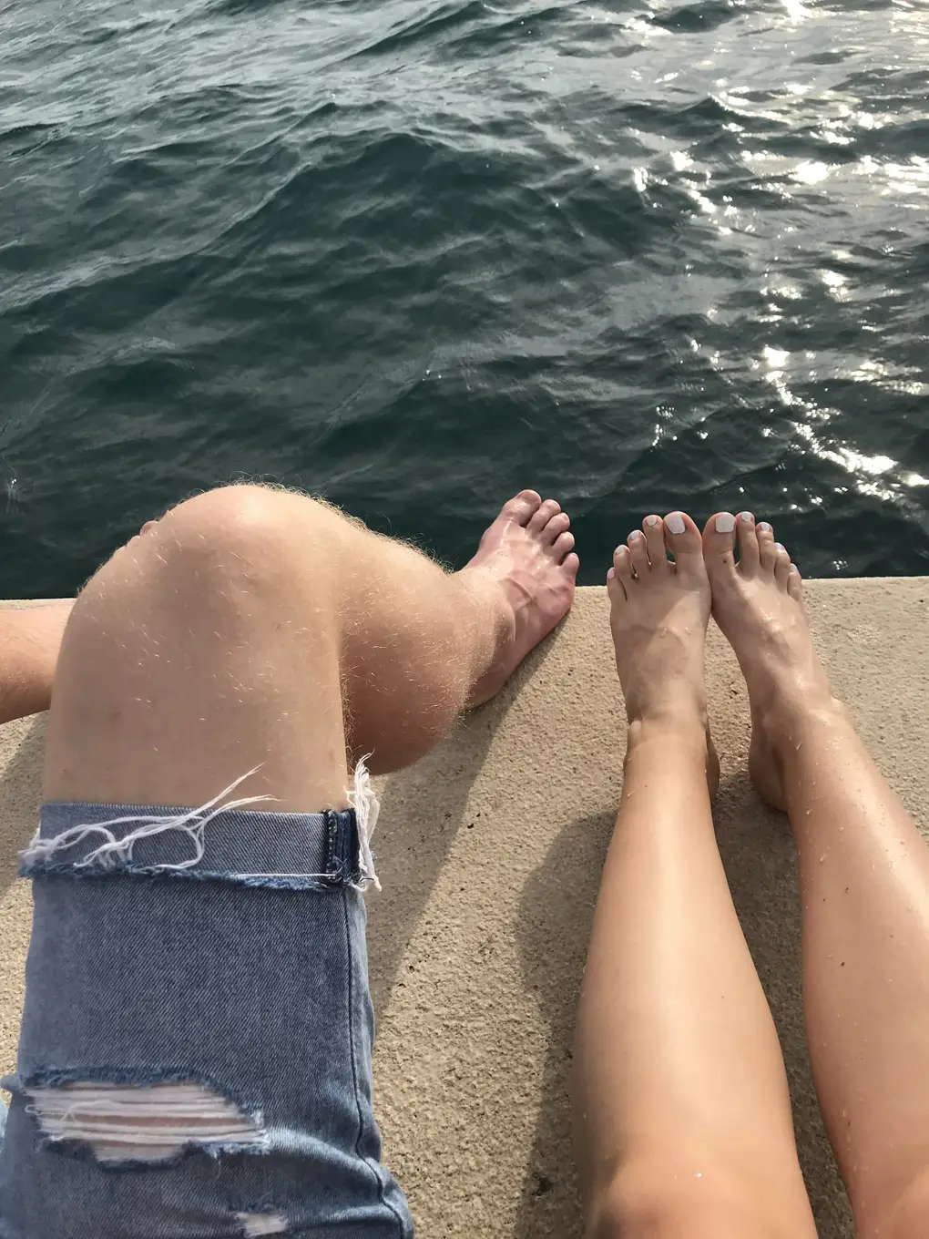 Von oben zwei Paar nasse Beine und Füße auf einer Steinkante über dem Meer, eines in zerrissenen Jeansshorts