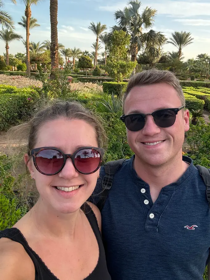 Selfie von Rebecca und Max mit Sonnenbrillen, hinter ihnen Palmen, Hecken und blühende Beete