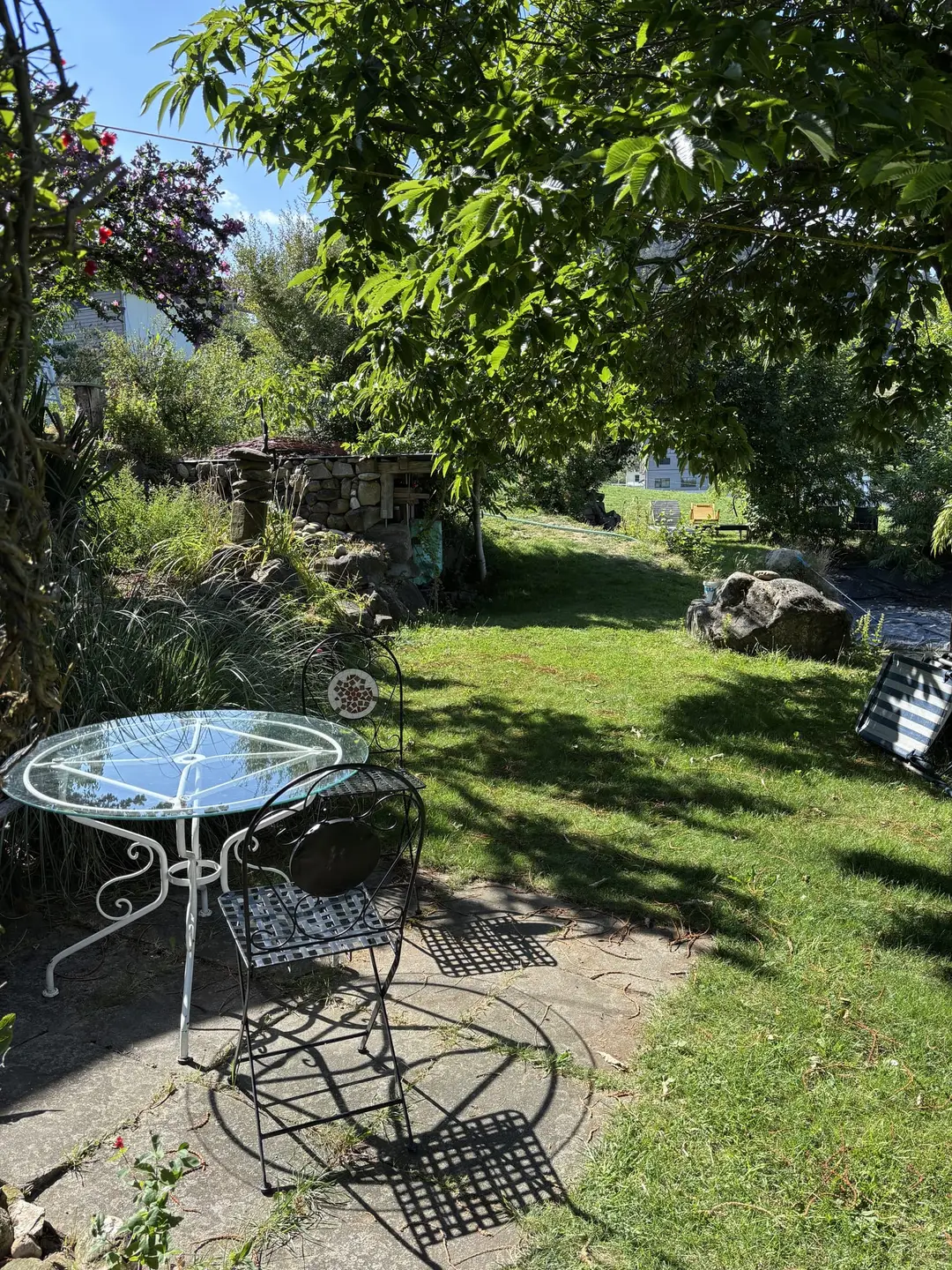 Ein Garten in der Morgensonne mit Rasen, Bäumen und Sträuchern, vorne ein runder Glastisch mit verschnörkelten Metallstühlen