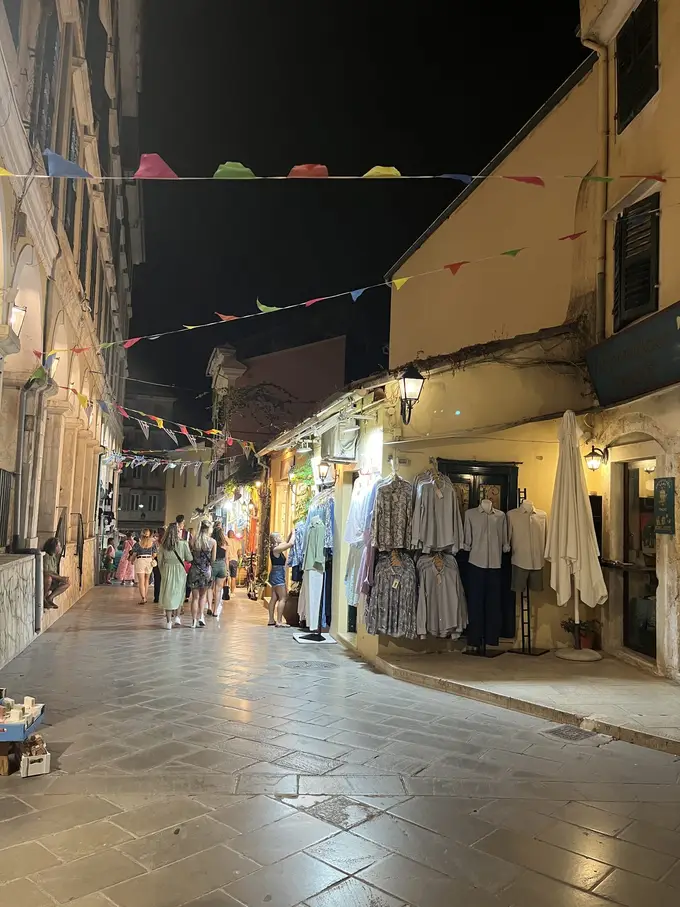 Beleuchtete Gasse bei Nacht mit Kleiderständern vor den Läden und Wimpeln über der Straße