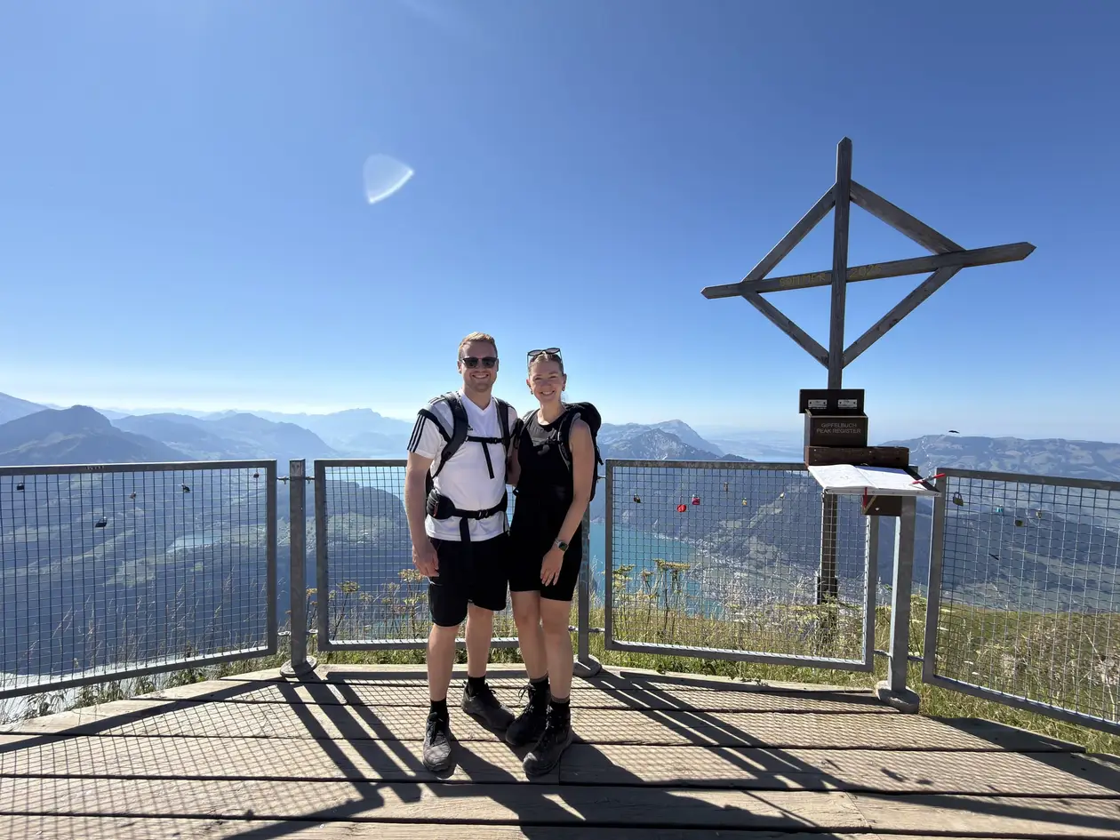 Max und Rebecca auf einer Aussichtsplattform neben einem hölzernen Gipfelkreuz, hinter ihnen Berge und ein See