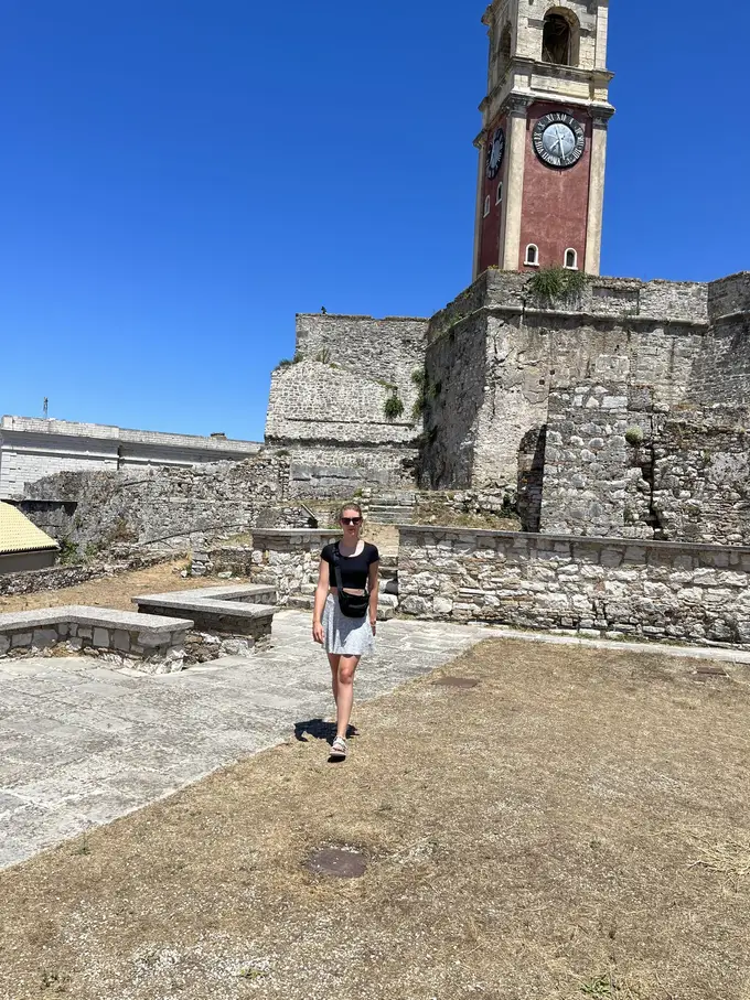 Rebecca geht über einen Platz in der Festung, dahinter ein roter Glockenturm auf alten Mauern